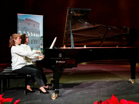 Two women in white blouses perform together at a black Steinway grand piano on a dimly lit stage, with an image of the Roman Colosseum behind them.