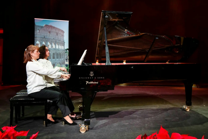 Two women in white blouses perform together at a black Steinway grand piano on a dimly lit stage, with an image of the Roman Colosseum behind them.