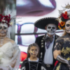 Two women wearing elaborate floral headdresses and painted skull makeup, one in a white bridal gown and the other in a black dress, pose with a child and a man in a sombrero.