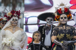 Two women wearing elaborate floral headdresses and painted skull makeup, one in a white bridal gown and the other in a black dress, pose with a child and a man in a sombrero.
