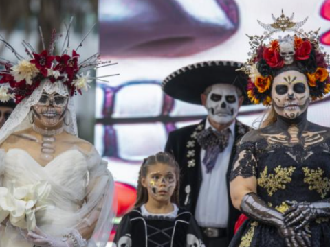 Two women wearing elaborate floral headdresses and painted skull makeup, one in a white bridal gown and the other in a black dress, pose with a child and a man in a sombrero.