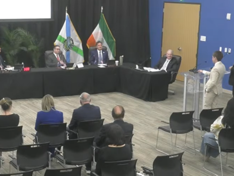 Wide view of a Coral Gables City Commission meeting, with commissioners seated behind black tables and two students in suits standing near a clear lectern at the right.