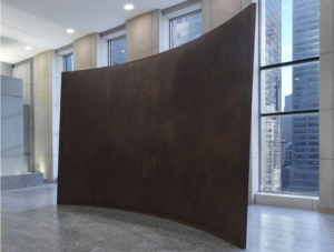 Richard Serra's Fin, a large, dark, curved steel wall with a sloping upper edge, displayed in a stone-floored Manhattan office lobby with floor-to-ceiling windows.