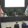 Coral Gables city commissioners sit behind a long black-draped table during an inspector general workshop, with several attendees seated in the foreground and flags behind the dais.