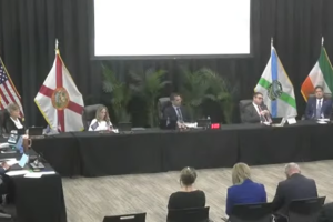 Coral Gables city commissioners sit behind a long black-draped table during an inspector general workshop, with several attendees seated in the foreground and flags behind the dais.