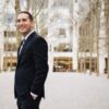 Pianist Mackenzie Melemed smiles while standing in a dark suit and tie in a tree-lined urban plaza with modern buildings behind him.