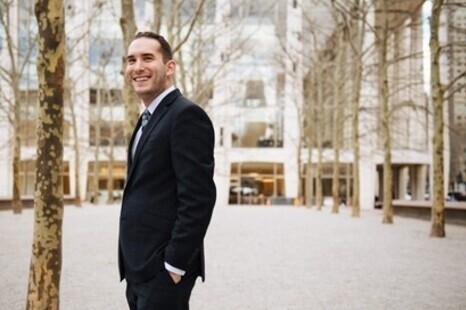 Pianist Mackenzie Melemed smiles while standing in a dark suit and tie in a tree-lined urban plaza with modern buildings behind him.
