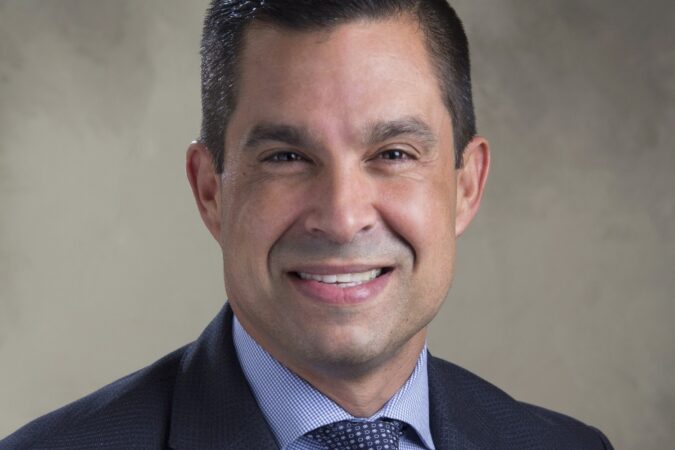 Head-and-shoulders portrait of Coral Gables Mayor Vince Lago smiling in a dark suit, blue checked shirt and patterned tie against a neutral background.