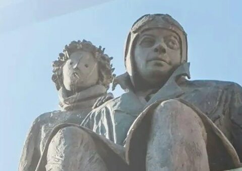 Sculpture of Antoine de Saint-Exupéry seated beside the Little Prince against a pale blue sky.