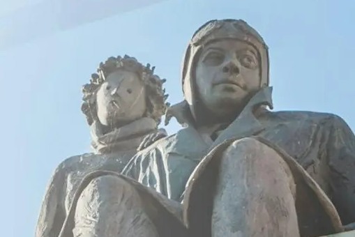 Sculpture of Antoine de Saint-Exupéry seated beside the Little Prince against a pale blue sky.