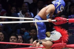 Two masked little-person Lucha Libre wrestlers compete in a wrestling ring as spectators watch from the background.