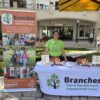 Woman in a bright green Branches T-shirt stands behind a table displaying Thanksgiving food donations, with a Branches banner and Shops at Merrick Park storefronts in the background.