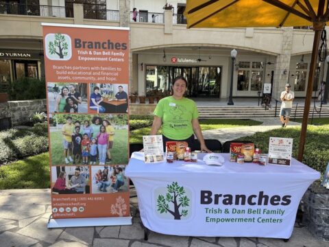 Woman in a bright green Branches T-shirt stands behind a table displaying Thanksgiving food donations, with a Branches banner and Shops at Merrick Park storefronts in the background.