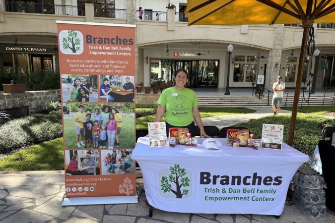 Woman in a bright green Branches T-shirt stands behind a table displaying Thanksgiving food donations, with a Branches banner and Shops at Merrick Park storefronts in the background.