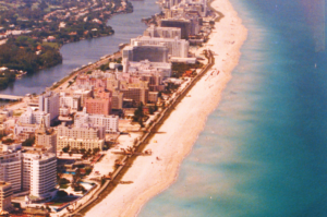 Aerial photograph of Miami Beach showing beachfront hotels, a broad sandy beach, turquoise Atlantic Ocean waters and an inland waterway.
