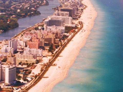 Aerial photograph of Miami Beach showing beachfront hotels, a broad sandy beach, turquoise Atlantic Ocean waters and an inland waterway.