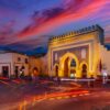Bab Bou Jeloud, Fez's historic Blue Gate, illuminated at dusk with three horseshoe arches, intricate blue mosaic tilework and battlements beneath a pink and purple sunset sky, with red traffic light trails in the foreground.