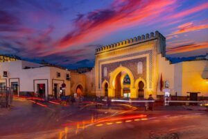 Bab Bou Jeloud, Fez's historic Blue Gate, illuminated at dusk with three horseshoe arches, intricate blue mosaic tilework and battlements beneath a pink and purple sunset sky, with red traffic light trails in the foreground.