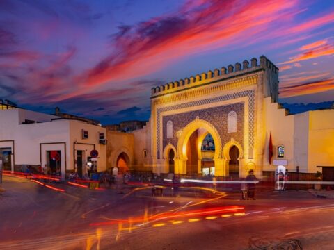 Bab Bou Jeloud, Fez's historic Blue Gate, illuminated at dusk with three horseshoe arches, intricate blue mosaic tilework and battlements beneath a pink and purple sunset sky, with red traffic light trails in the foreground.