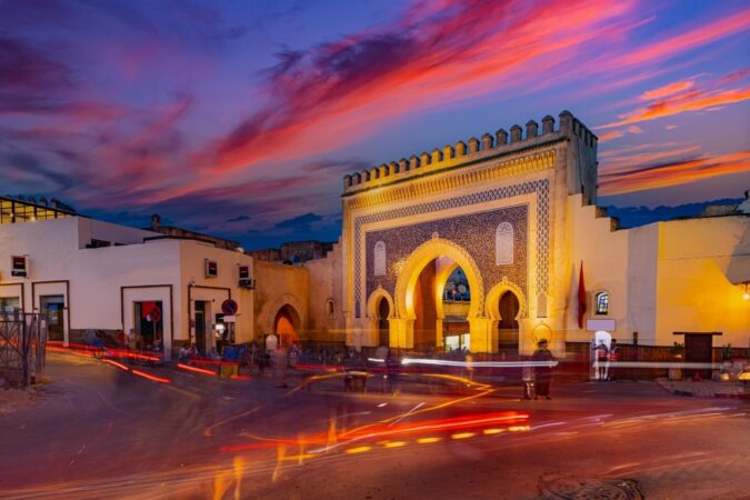 Bab Bou Jeloud, Fez's historic Blue Gate, illuminated at dusk with three horseshoe arches, intricate blue mosaic tilework and battlements beneath a pink and purple sunset sky, with red traffic light trails in the foreground.