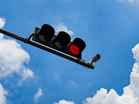 Red traffic signal and mounted red-light enforcement camera against a blue sky with clouds.
