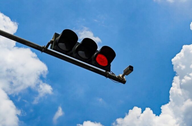 Red traffic signal and mounted red-light enforcement camera against a blue sky with clouds.