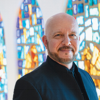 Dr. James K. Bass, artistic director of Seraphic Fire, wearing a black jacket and standing before colorful blue, gold and white stained-glass windows.