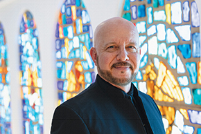 Dr. James K. Bass, artistic director of Seraphic Fire, wearing a black jacket and standing before colorful blue, gold and white stained-glass windows.