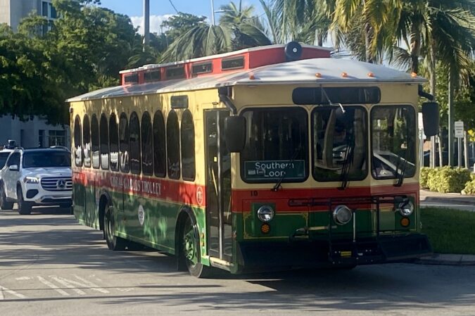 Coral Gables Southern Loop trolley with red, green and cream exterior traveling along a palm-lined street.