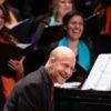 Shawn Kirchner smiles while seated at a grand piano, with choral singers behind him holding music folders during a performance.