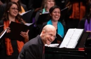 Shawn Kirchner smiles while seated at a grand piano, with choral singers behind him holding music folders during a performance.