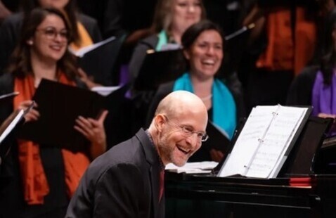 Shawn Kirchner smiles while seated at a grand piano, with choral singers behind him holding music folders during a performance.